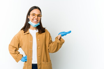 Young latin woman wearing a mask to protect from covid isolated on white background showing a copy space on a palm and holding another hand on waist.