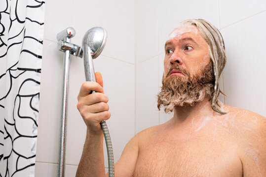 Confused bearded man with soapy head standing in the bathroom and looking at the shower while the water supply has stopped. No water coming from tap.