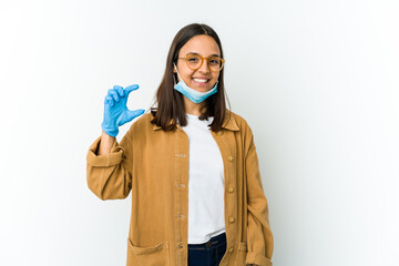 Young latin woman wearing a mask to protect from covid isolated on white background holding something little with forefingers, smiling and confident.