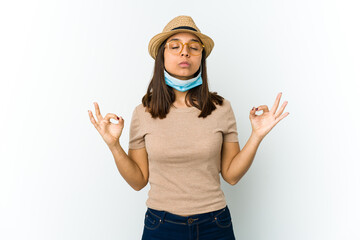 Young latin woman wearing hat and mask to protect from covid isolated on white background relaxes after hard working day, she is performing yoga.