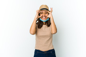 Young latin woman wearing hat and mask to protect from covid isolated on white background showing okay sign over eyes