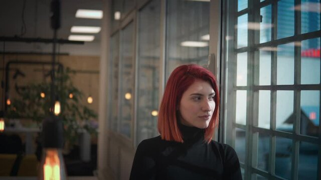 Pretty thoughtful lonely businesswoman with red hair looking out a window in modern office
