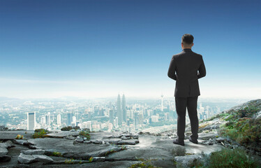 Back view of young businessman standing on mountain top looking at cityscape-Leadership concept