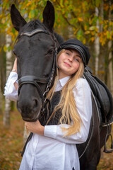 Obraz premium portrait of a girl with blonde hair , wearing a cap, and a Bay horse on the background of a forest. 