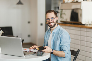 Smiling man in glasses sit at table in coffee shop cafe restaurant indoors working studying on laptop computer pay off with cell phone bank payment terminal. Freelance mobile office business concept.