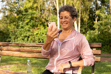 Old aged brazilian woman looking mobile screen to use app. Outdoors in sunny day. Network, messaging, internet, lifestyle concept.