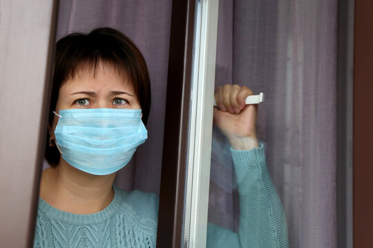 Sick woman in a face medical mask looks out of the window. Coronavirus quarantine during the covid-19 pandemic