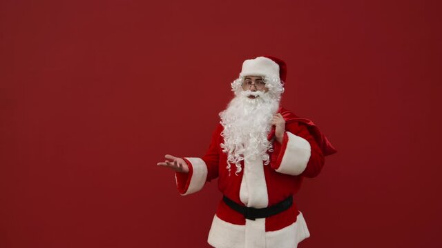 Confused man in a suite suit stands on a red background with a bag of gifts on his back and looks around in confusion. Puzzled Santa at Christmas.