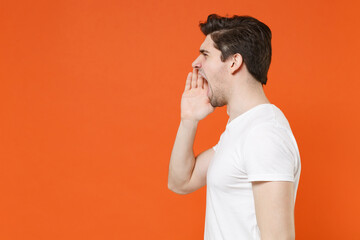 Side view of crazy young man wearing basic casual blank empty white t-shirt standing screaming with hands gesture near mouth looking aside isolated on bright orange colour background, studio portrait.