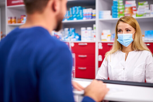 Young Female Apothecary In Protective Medical Mask And Young Caucasian Man Customer Buying Drug At Drugstore. Medicine, Consumerism Concept. During Coronavirus Epidemic