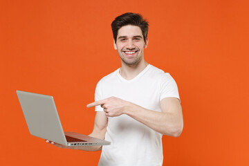 Smiling handsome young man in basic blank white t-shirt standing working pointing index finger on laptop pc computer looking camera isolated on bright orange colour wall background, studio portrait.
