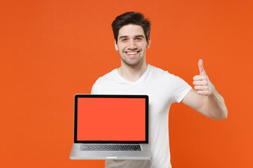 Smiling cheerful handsome young man 20s wearing basic casual white t-shirt hold laptop pc computer with blank empty screen showing thumb up isolated on bright orange colour background studio portrait.