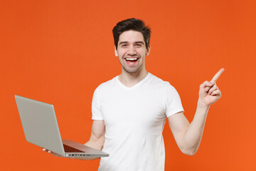 Laughing young man in basic casual blank white t-shirt standing working on laptop pc computer pointing index finger aside up looking camera isolated on bright orange colour background studio portrait.