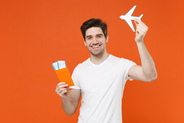 Smiling funny young traveler tourist man 20s wearing white t-shirt hold passport ticket air plane isolated on orange background. Passenger traveling on weekend getaway. Air flight journey concept.