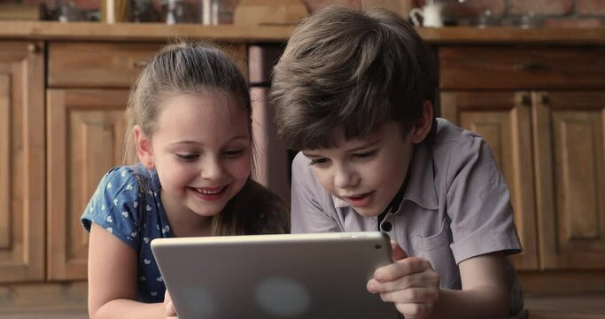 Close up little boy and girl lying on wooden warm floor at home kitchen with tablet device, watch cartoons, online video. Children development using modern technology, addicted kids, bad habit concept
