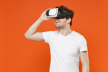 Smiling cheerful funny handsome young man 20s wearing basic casual white blank empty t-shirt standing watching in vr headset gadget isolated on bright orange colour background studio portrait.
