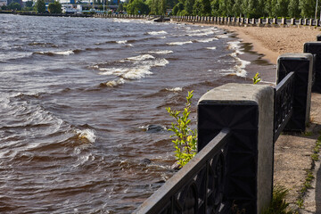 Panorama of the embankment by the lake.Along the sandy shore of the lake, there is a granite embankment, which is fenced with a cast-iron fence with pedestals. On the lake, the wind raised waves. 