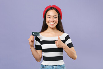 Naklejka premium Smiling funny young brunette asian woman 20s wearing striped t-shirt red beret standing holding in hand credit bank card showing thumb up isolated on pastel violet colour background studio portrait.