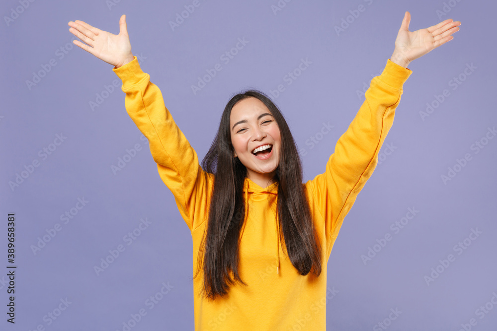 Wall mural Joyful cheerful laughing attractive young brunette asian woman in casual basic yellow hoodie standing rising spreading hands looking camera isolated on pastel violet colour background studio portrait. - Wall murals
