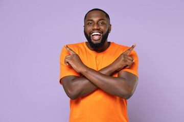 Excited young african american man in basic casual orange t-shirt holding hands crossed pointing index fingers aside on mock up copy space isolated on pastel violet colour background studio portrait.