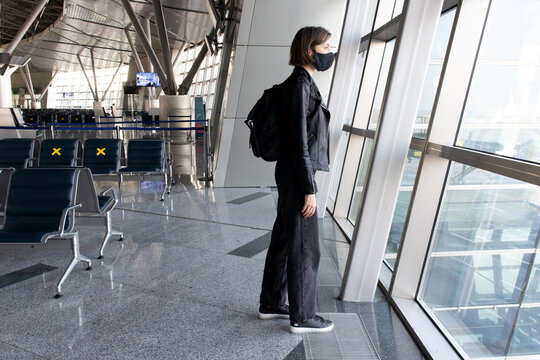Young woman in a protective black mask with a backpack near window at the international airport terminal. Traveling during the coronavirus pandemic