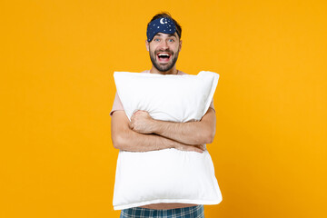 Excited cheerful young bearded man 20s in pajamas home wear sleep mask hold pillow while resting at home isolated on bright yellow colour background studio portrait. Relax good mood lifestyle concept.
