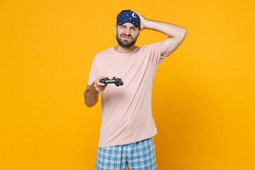 Displeased young man in pajamas home wear sleep mask play pc game with joystick console put hand on...