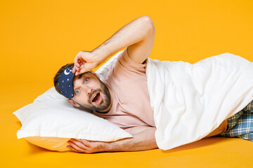 Excited surprised shocked young bearded man in pajamas home wear sleep mask lying with pillow blanket isolated on bright yellow colour background studio portrait. Relax good mood lifestyle concept.