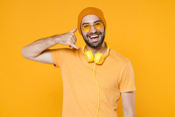 Cheerful young bearded man 20s wearing basic casual t-shirt headphones eyeglasses hat standing doing phone gesture like says call me back isolated on bright yellow colour background, studio portrait.