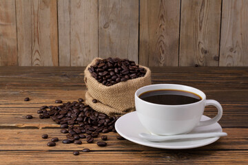 White coffee cup with dark black coffee, with saucer and spoon, fragrant roasted coffee beans in a bag placed on an old wooden floor.