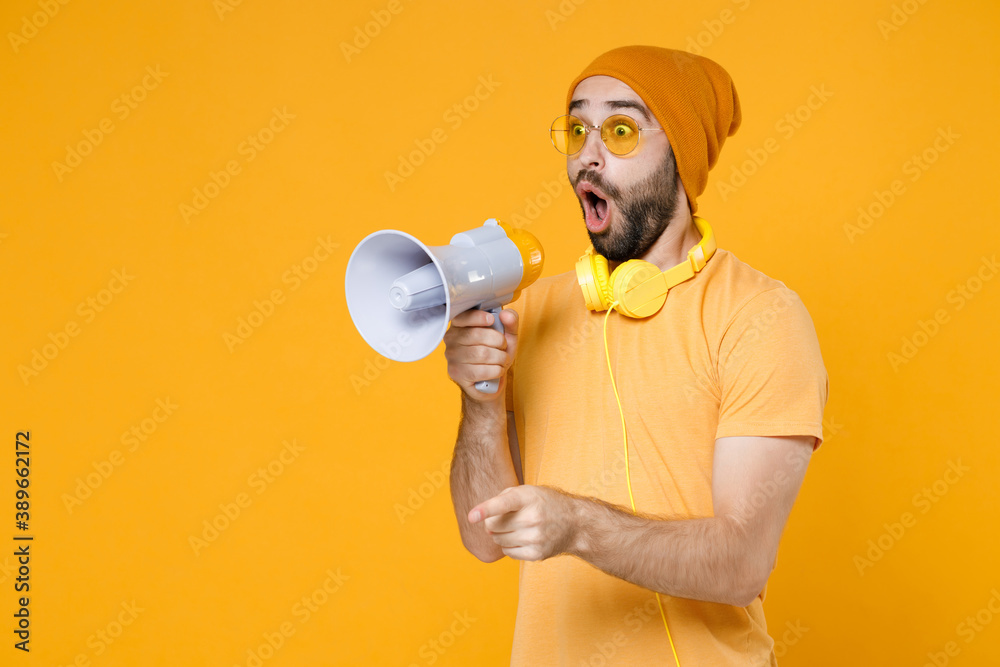 Wall mural Shocked amazed young bearded man 20s wearing basic casual t-shirt headphones eyeglasses hat standing screaming in megaphone pointing index finger aside isolated on yellow background, studio portrait.