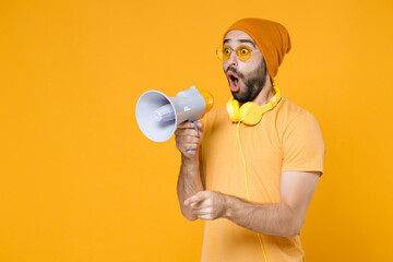 Shocked amazed young bearded man 20s wearing basic casual t-shirt headphones eyeglasses hat standing screaming in megaphone pointing index finger aside isolated on yellow background, studio portrait.