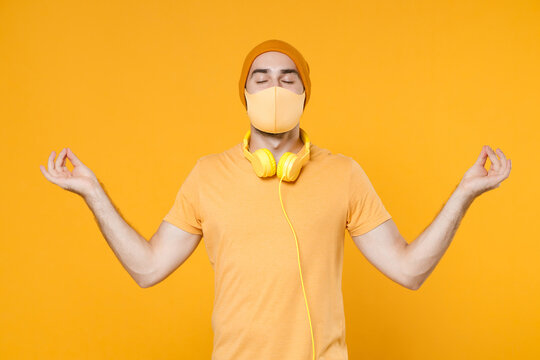Young man in t-shirt headphones face mask safe from coronavirus virus covid-19 during pandemic quarantine hold hands in yoga gesture, relaxing meditating isolated on yellow background studio portrait.