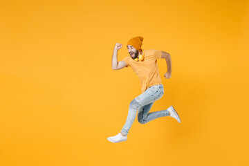 Fototapeta na wymiar Full length side view of cheerful crazy screaming young man wearing basic casual t-shirt headphones hat jumping like running looking aside isolated on bright yellow colour background, studio portrait.