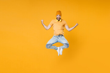 Obraz premium Full length of overjoyed screaming young man 20s wearing basic casual t-shirt headphones hat jumping doing winner gesture looking camera isolated on bright yellow colour background, studio portrait.