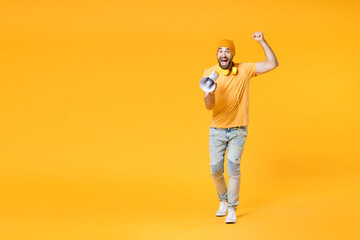 Fototapeta na wymiar Full length of shocked amazed young man wearing basic casual t-shirt headphones hat screaming in megaphone clenching fist looking camera isolated on bright yellow colour background, studio portrait.