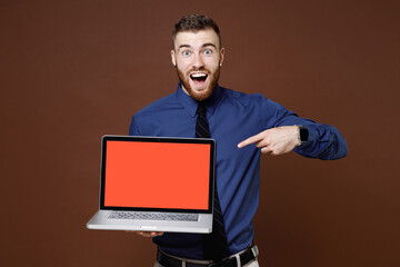 Shocked young business man in blue shirt tie point index finger on laptop pc computer with blank empty screen isolated on brown background studio portrait. Achievement career wealth business concept.