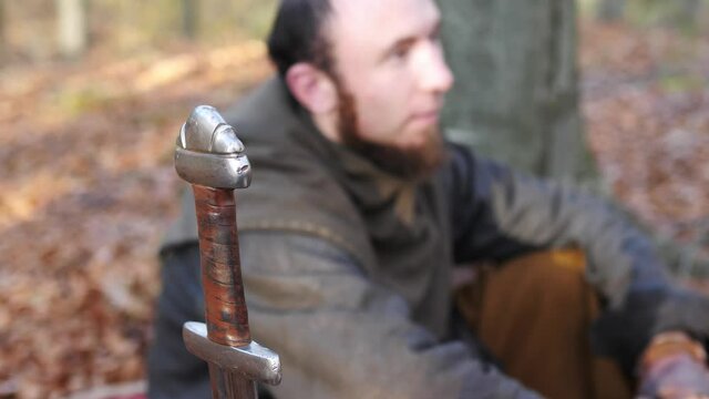 Viking warriors sit in the forest and talk in the campground, while the sun shines through the foliage. Medieval reconstruction.