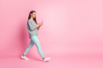 Full size profile photo of optimistic cute young woman going look telephone wear blue jeans sneakers shirt isolated on pink color background