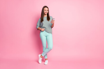 Full size photo of cheerful nice brunette hair woman stand look telephone wear blue trousers sneakers pullover isolated on pink background