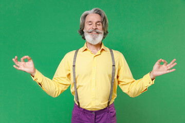 Smiling elderly gray-haired mustache bearded man in yellow shirt suspenders hold hands in yoga gesture, relaxing meditating, trying to calm down isolated on green colour background, studio portrait.