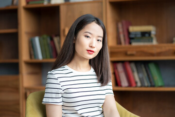 Portrait of beautiful young Asian woman in striped t shirt sitting in chair against background of bookcases in library or business office