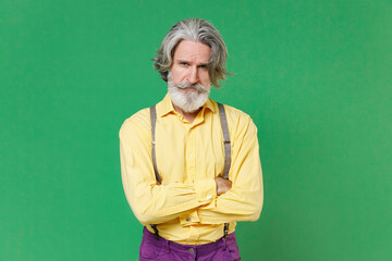 Displeased dissatisfied elderly gray-haired mustache bearded man wearing casual yellow shirt suspenders holding hands crossed looking camera isolated on bright green colour background studio portrait.