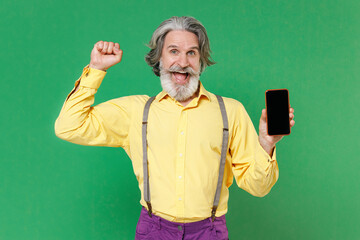 Excited elderly gray-haired mustache man in yellow shirt suspenders hold mobile cell phone with blank empty screen mock up copy space doing winner gesture isolated on green background studio portrait.
