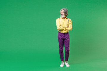 Full length of smiling elderly gray-haired mustache bearded man wearing casual yellow shirt suspenders holding hands crossed looking aside isolated on bright green colour background, studio portrait.