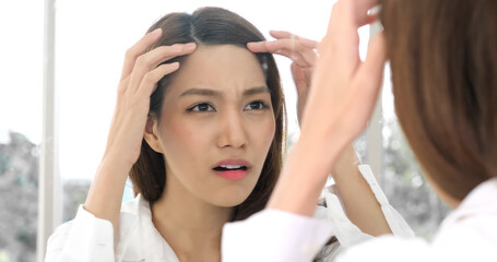 Portrait of worried young Asian woman looking in mirror, examining acne and wrinkles on her face. Women never applied facial cream Until face has wrinkles and acne. - Powered by Adobe