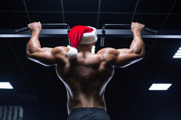 Fototapeta na wymiar Young sexy Santa Claus in a Christmas hat is working out pumping up back muscles in the gym gaining weight on machines