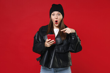 Shocked amazed young brunette woman 20s wearing casual black leather jacket white t-shirt hat pointing index finger on mobile cell phone typing sms message isolated on red background studio portrait.