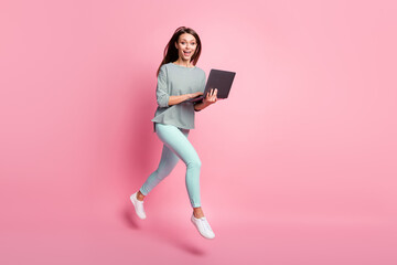Full size profile photo of optimistic funny brunette hair girl run jump typing laptop wear shirt pants sneakers isolated on pink background