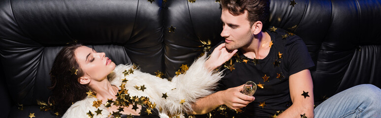 Man looking at passionate woman with glass of champagne lying on sofa, while confetti falling in...